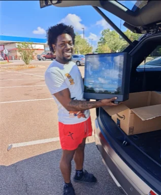 A smiling customer lifting a monitor out of an SUV cargo area, beside the shipping box.