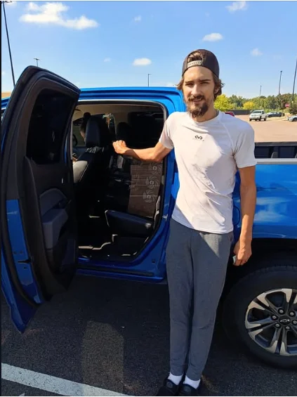 A customer standing at the open rear door of a blue pickup, the boxed PC on the back seat.